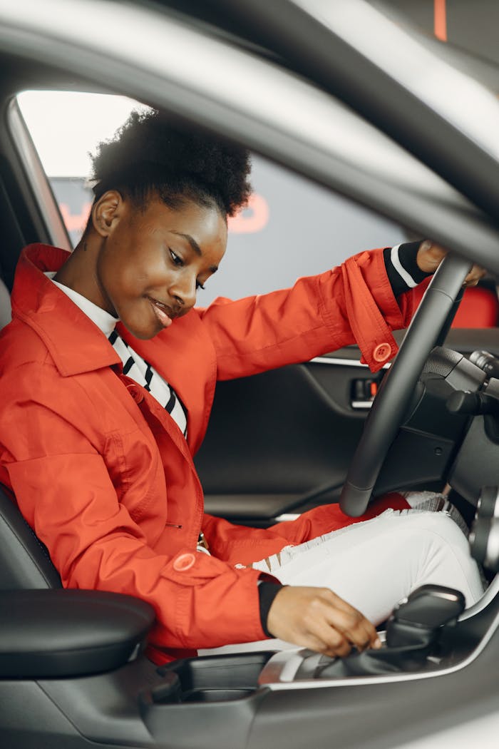 A cheerful woman in a red jacket adjusts car controls while seated inside a vehicle.